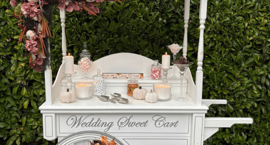 Our white candy cart with Wedding Sweet Cart painted on the front, set with jars of sweets, candles and autumn flowers against a hedge
