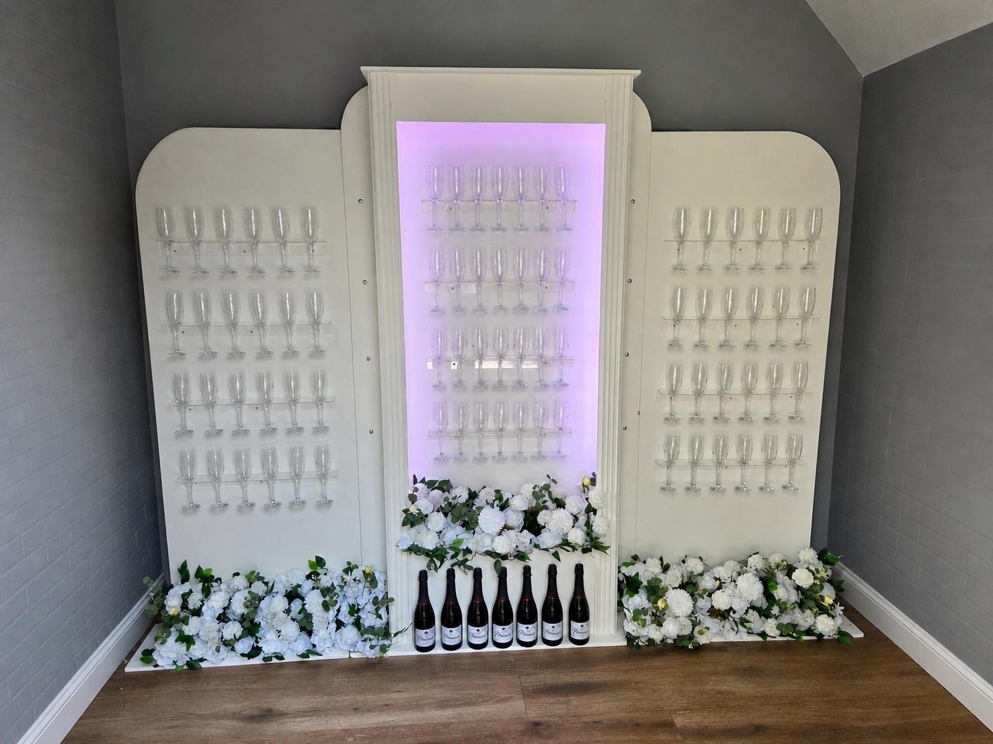 Our large three-panel prosecco wall with a lilac-lit centre panel, rows of empty flutes ready to fill and white flowers along its base