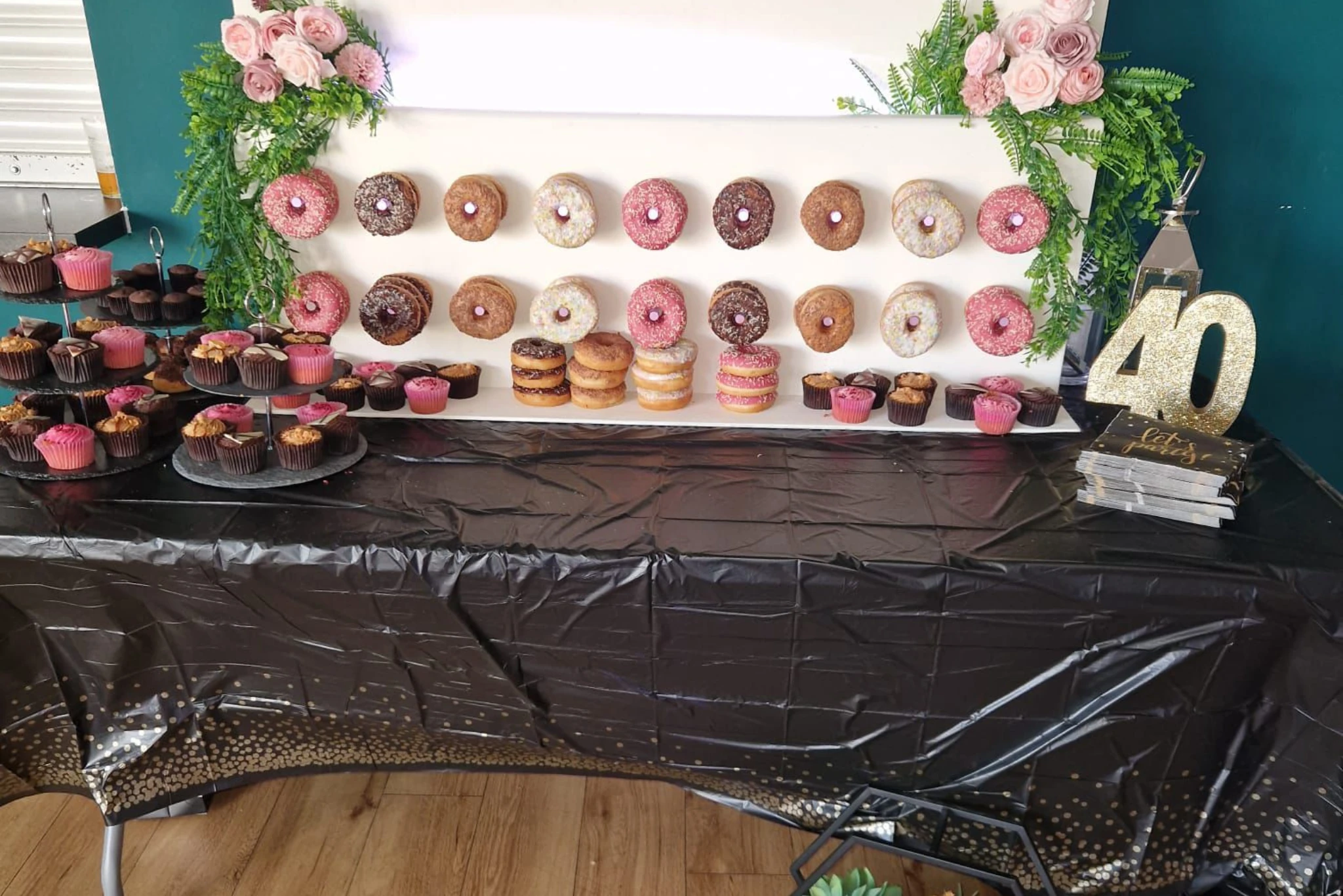 Our doughnut wall with two rows of iced doughnuts on pegs, pink roses and greenery at the corners, cupcakes on stands along the table and a gold glitter 40 beside it